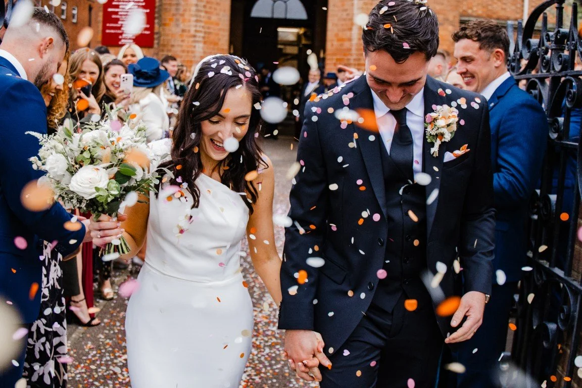 Bride and groom holding hands, walking through confetti-filled outdoor celebration, surrounded by smiling guests. The bride is in a white dress holding a bouquet, and the groom in a black suit with a boutonniere. The scene has a joyful, festive atmos