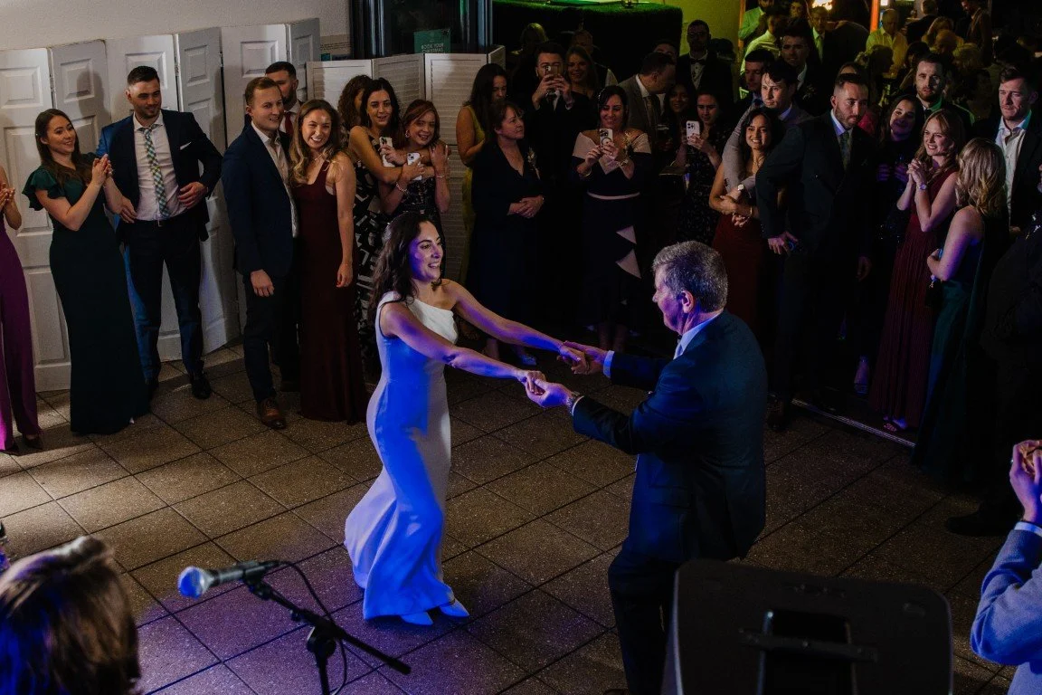 A bride and groom dancing at their wedding reception with friends and family watching and taking photos.