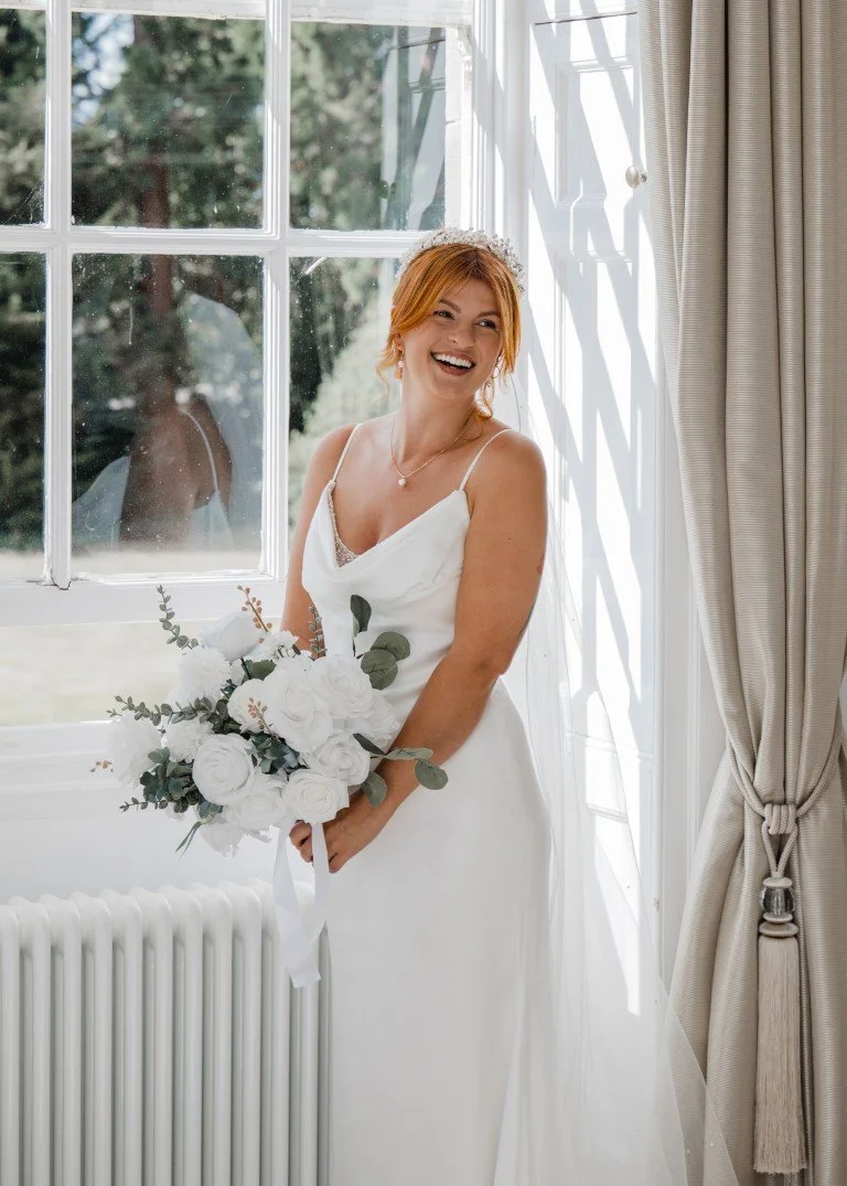 A bride in a white wedding dress holding a bouquet of white flowers and greenery, standing in front of a window with curtains, smiling and looking joyful.