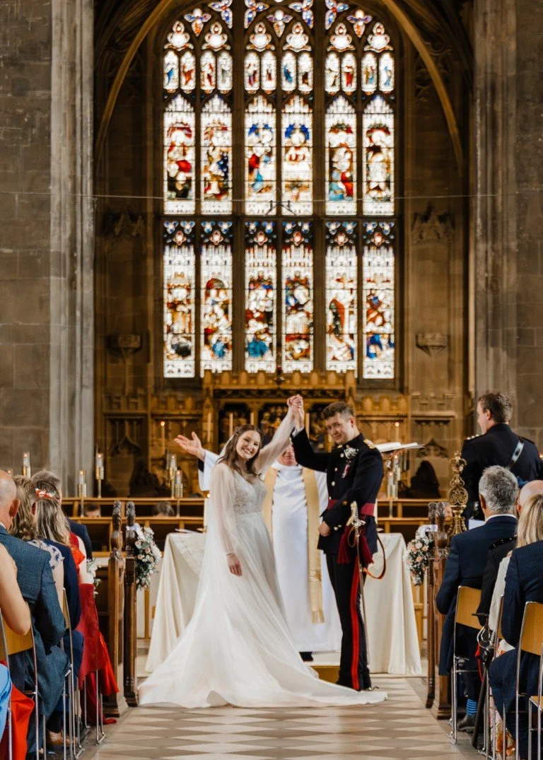 A bride and groom holding hands and smiling during their wedding ceremony inside a church with stained glass windows. The bride wears a white wedding dress, and the groom is in a formal military uniform. Guests are seated on either side of the aisle,