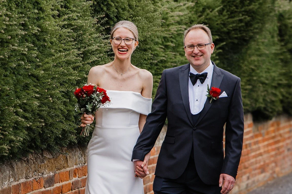 Happy couple dressed in wedding attire holding hands and walking outdoors on a brick sidewalk, with green bushes in the background.