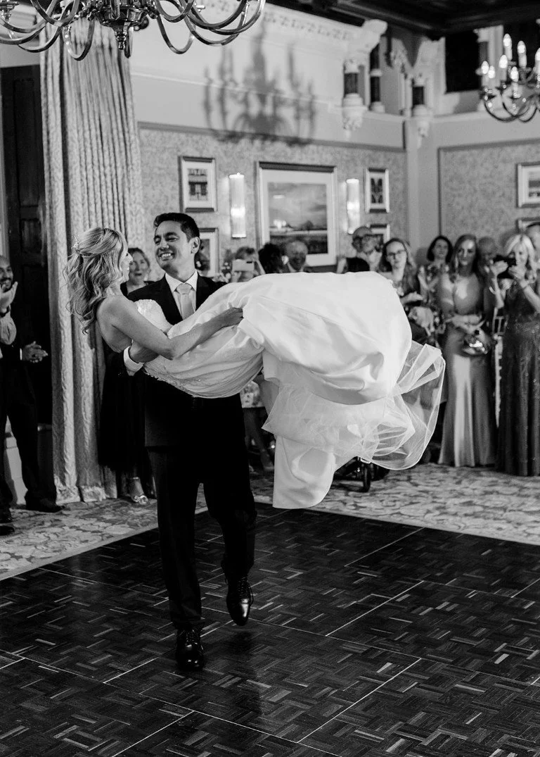 A groom is holding and spinning a bride during their wedding dance at a reception, with guests watching and smiling in the background.
