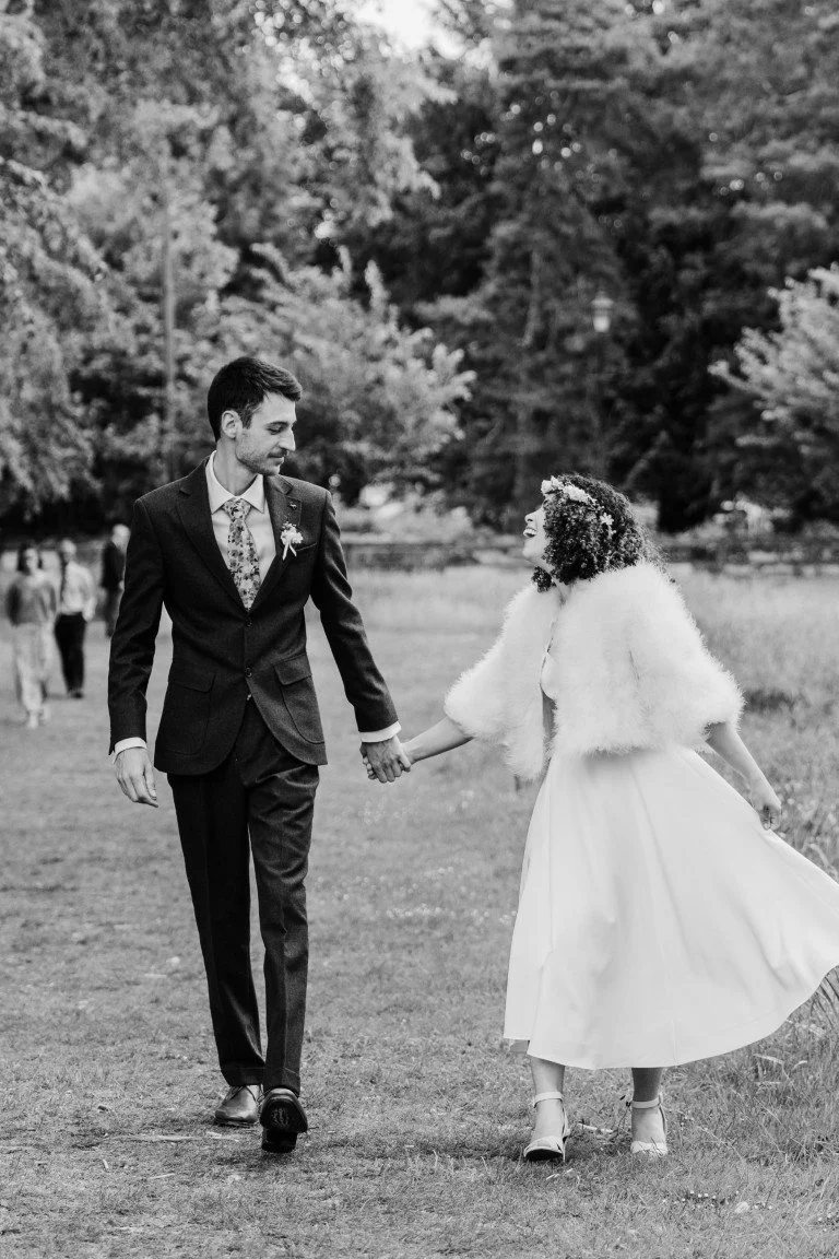 A black and white photo of a wedding couple holding hands and walking outdoors on a grassy area with trees in the background, with a few people blurred in the distance.