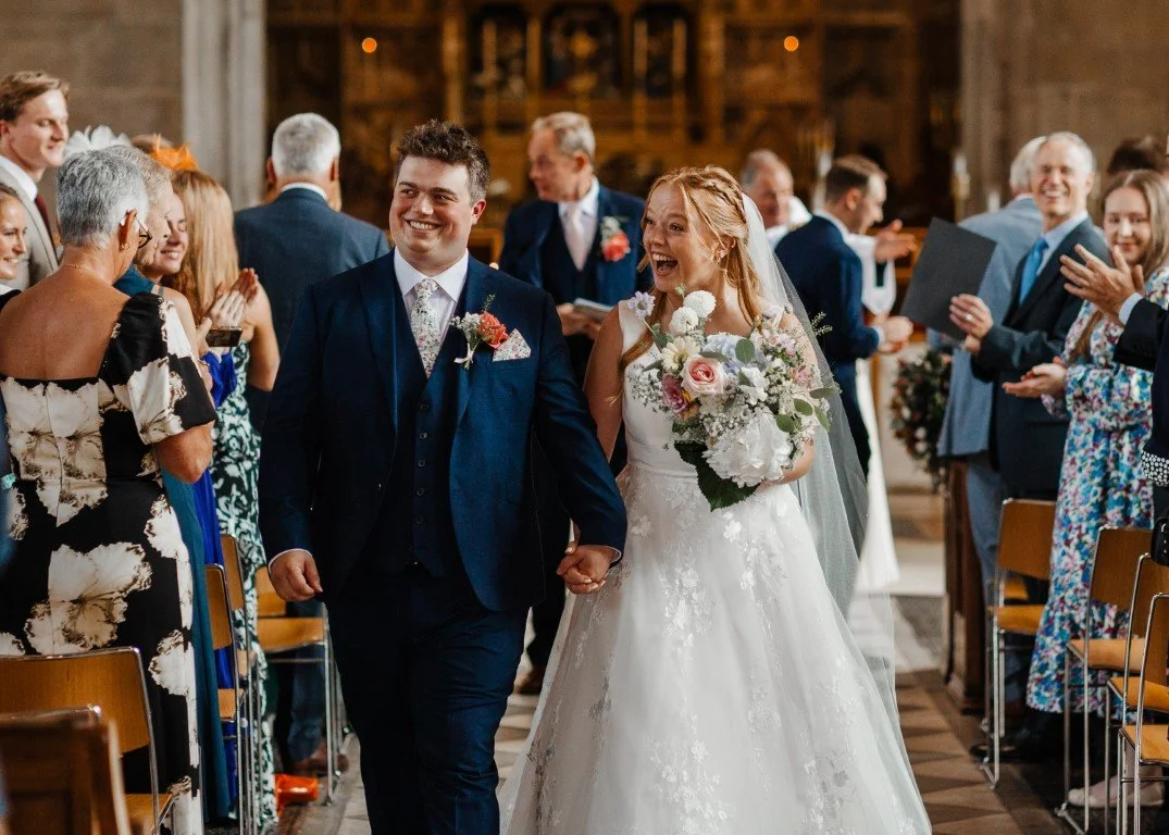 Bride and groom walking down the aisle hand in hand, smiling, as guests applaud and take photos in a church setting.