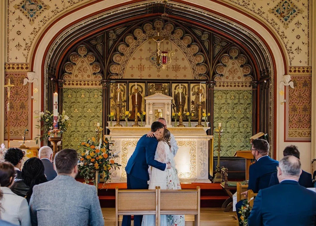 A wedding ceremony inside a church with a bride and groom embracing at the altar, surrounded by seated guests. The ornate altar features candles and floral arrangements.