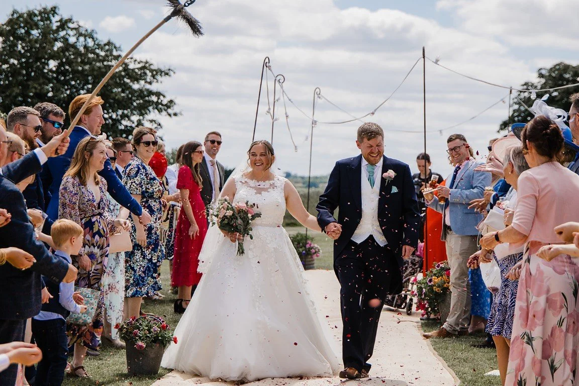 A bride and groom walking down the aisle, holding hands, smiling, surrounded by celebrating guests throwing confetti at an outdoor wedding ceremony.