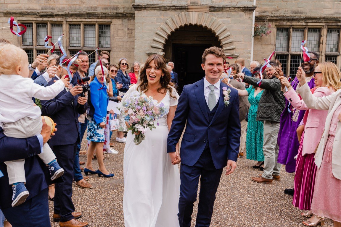 A newlywed couple walks hand in hand through a crowd of celebrating guests outside a historic stone building.