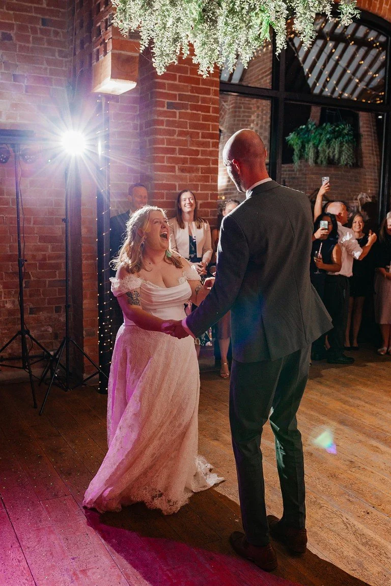 A bride and groom hold hands and dance during their wedding reception, with wedding guests smiling and taking photos in the background inside a decorated venue with brick walls and green plants.
