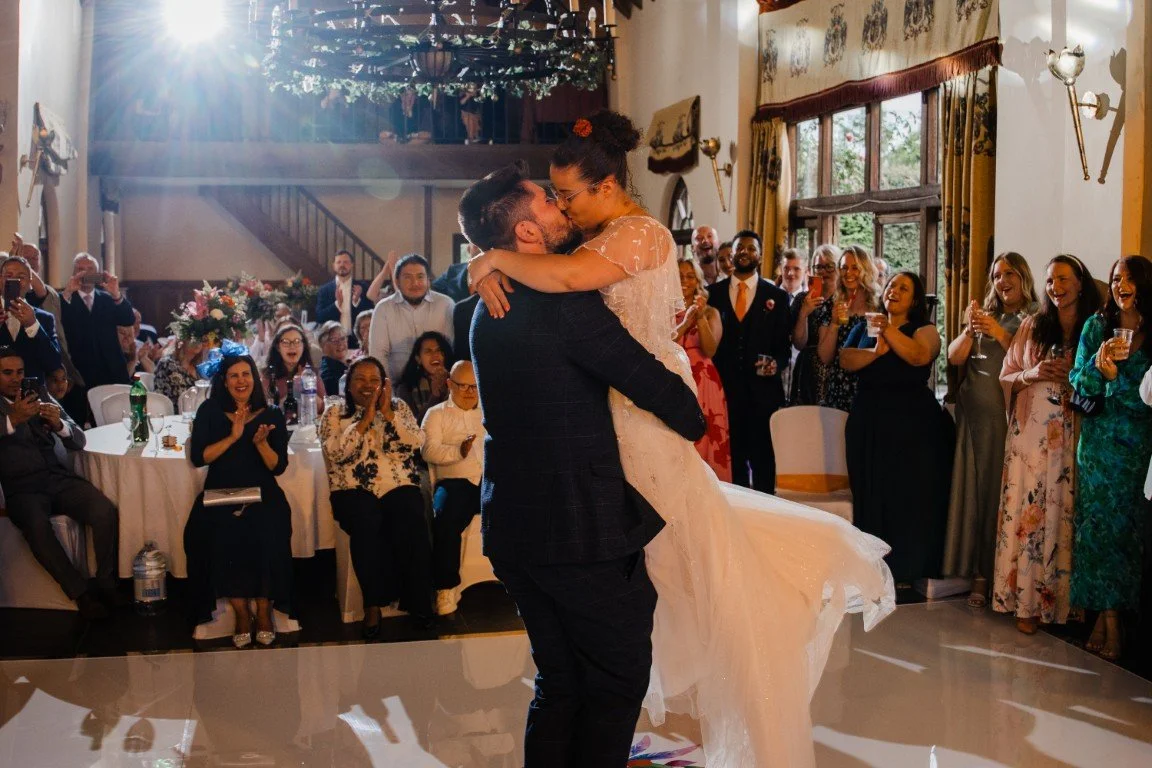 A newlywed couple sharing a kiss, with the groom lifting the bride during their wedding dance, while guests look on and celebrate in a decorated indoor venue.