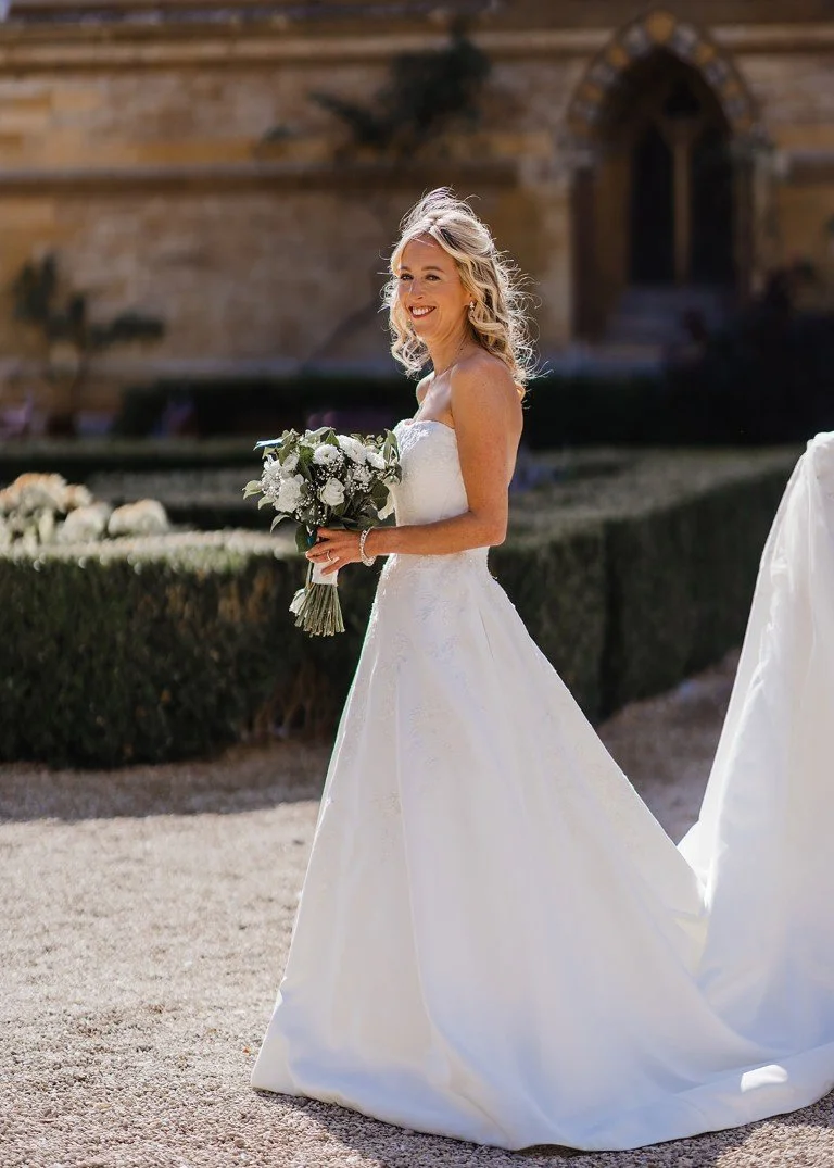 Bride in a white wedding dress holding a bouquet of white flowers, smiling outdoors near a historic building.
