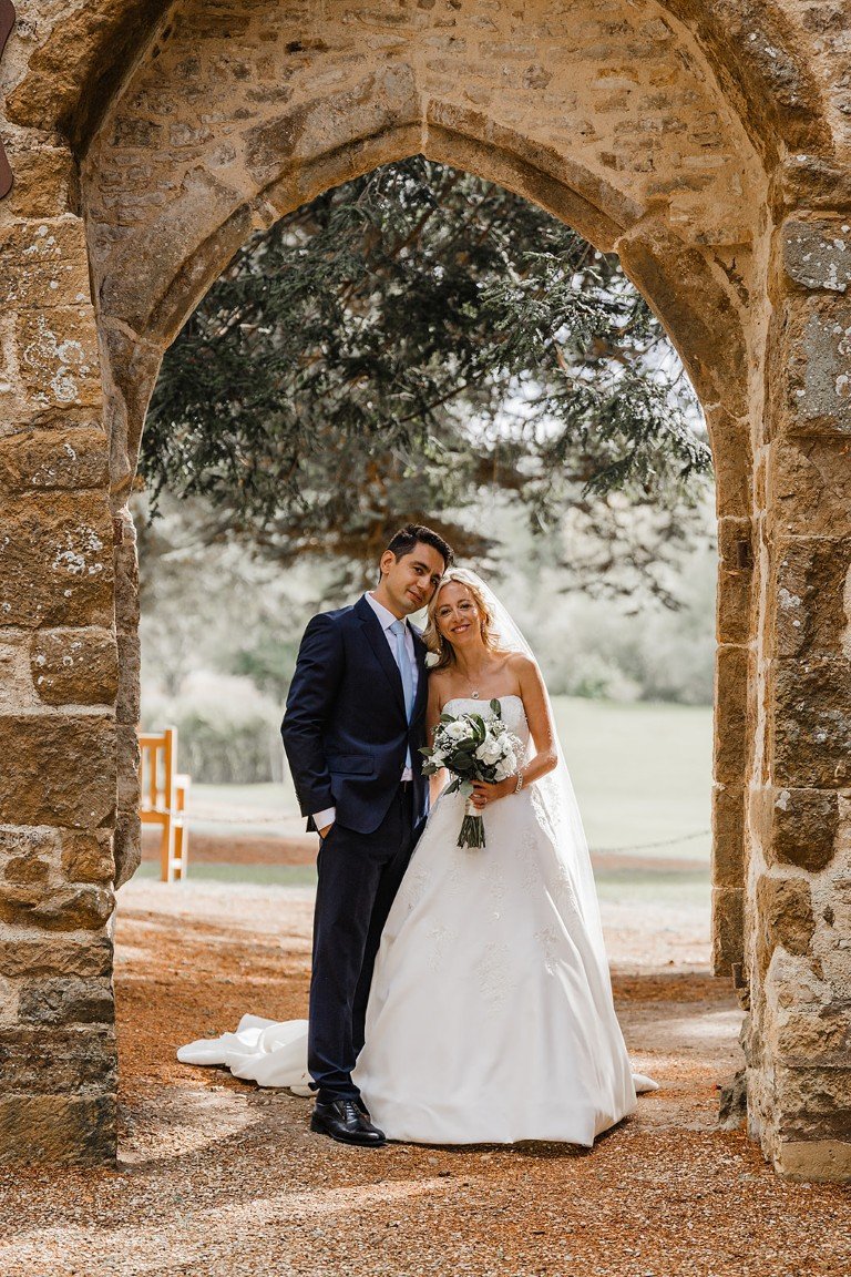 A newlywed couple stands under a stone archway outdoors, with the bride holding a bouquet of white flowers and the groom wearing a dark suit.