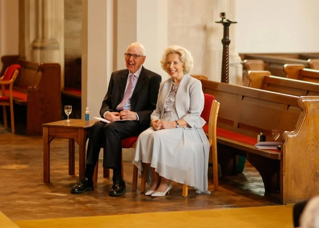 An elderly couple sitting on red-cushioned chairs inside a church, smiling and laughing during a formal event.