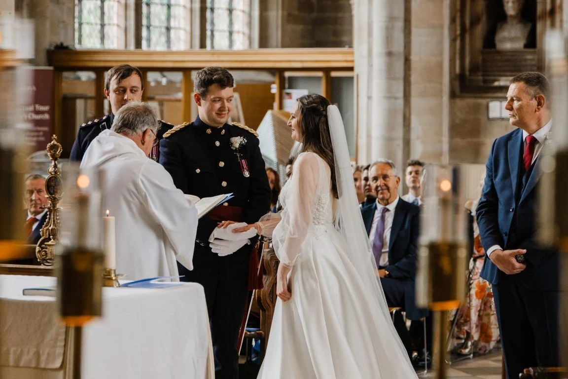 A wedding ceremony taking place inside a church, with a bride and groom standing before a priest, who is reading from a book, surrounded by guests and attendants.