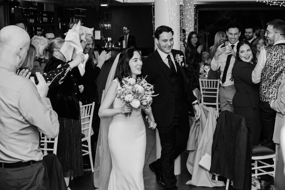 Black and white photo of a wedding reception with a bride and groom walking together through guests who are clapping and taking photos, surrounded by friends and family, in a decorated venue.