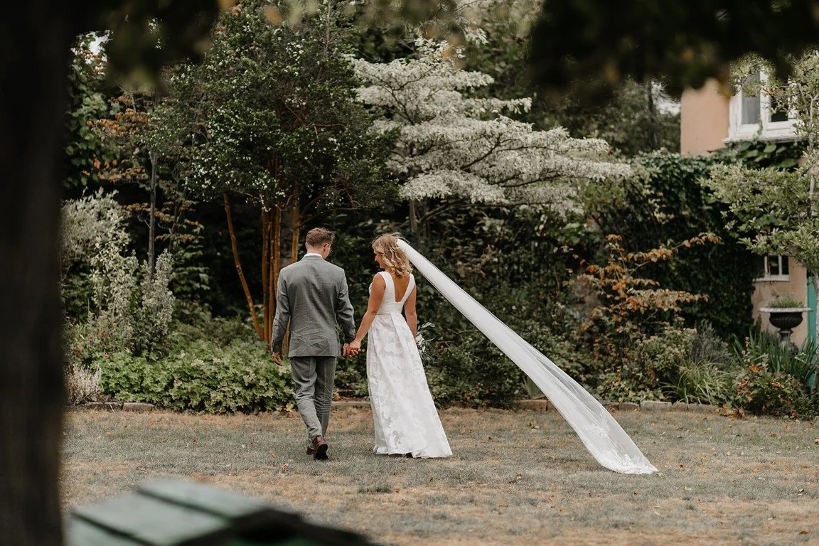 A bride and groom walking hand in hand in a garden, surrounded by greenery and flowering trees, with the bride wearing a white wedding dress and a long veil.