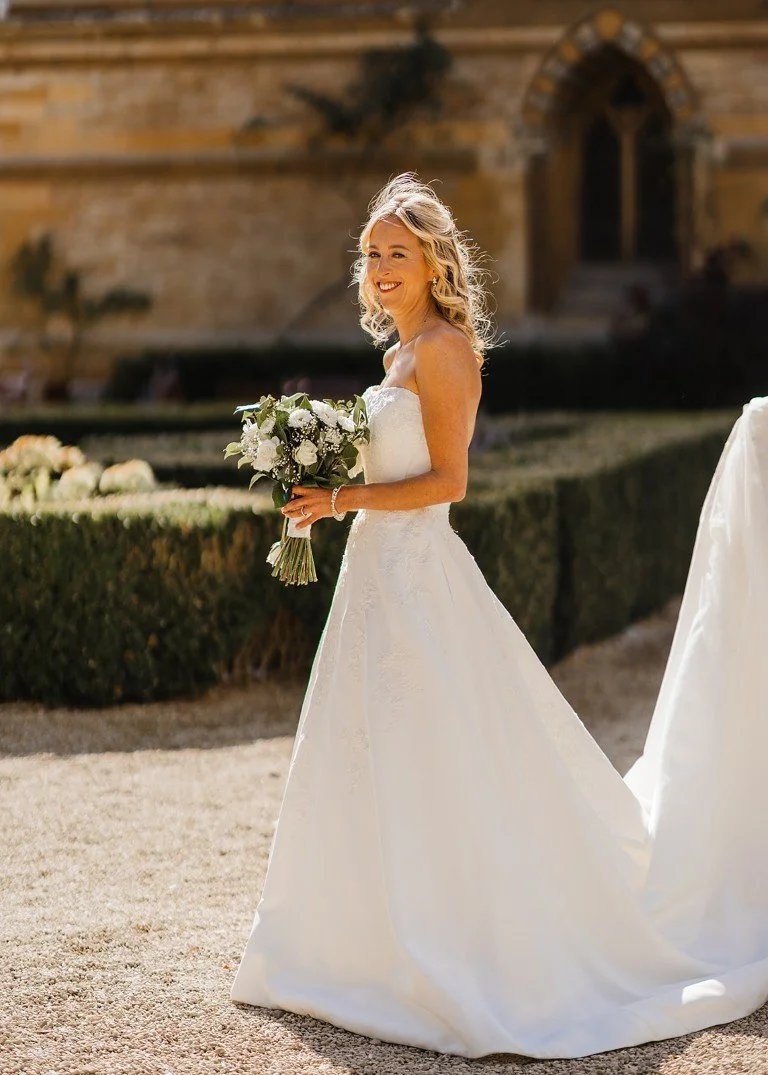 A smiling bride in a white wedding dress holding a bouquet of white flowers, standing outdoors on a sunny day with a stone building and greenery in the background.