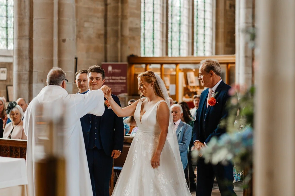 A wedding ceremony inside a church with a bride and groom standing in front of a priest. The bride is wearing a white gown and veil, and the groom is in a navy suit. They are holding hands, and guests are seated in the background.