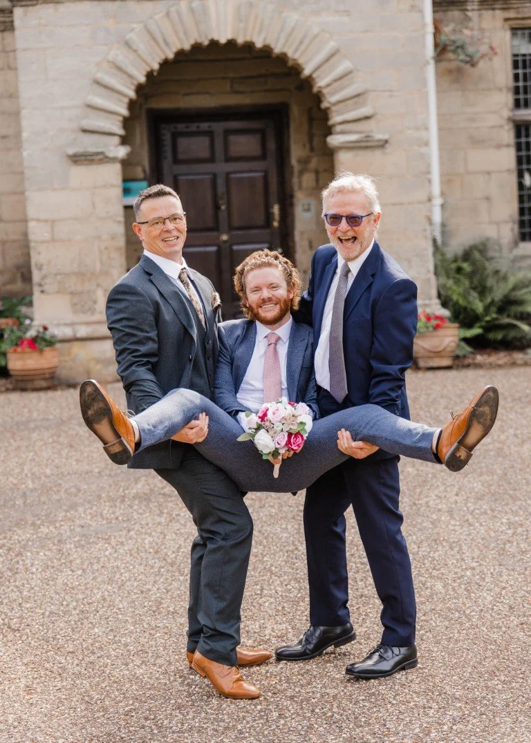 Three men dressed in formal suits standing outside a stone building, with two men lifting another man in a celebration pose. The man being lifted is holding a bouquet of flowers and smiling. The other two men are smiling and wearing sunglasses.