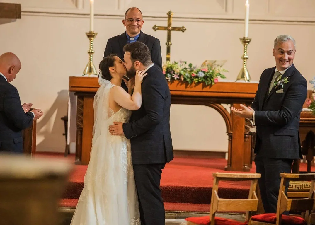 A couple in wedding attire kiss at the altar of a church during their wedding ceremony. The bride is in a white gown and veil, and the groom in a dark suit. A priest stands behind them, and another man in a suit is clapping on the right. The backgrou
