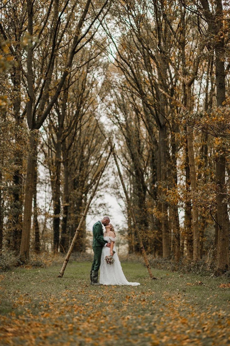 A couple dressed in wedding attire sharing a kiss under a teepee made of tall, leaning tree branches in a forest with autumn leaves.
