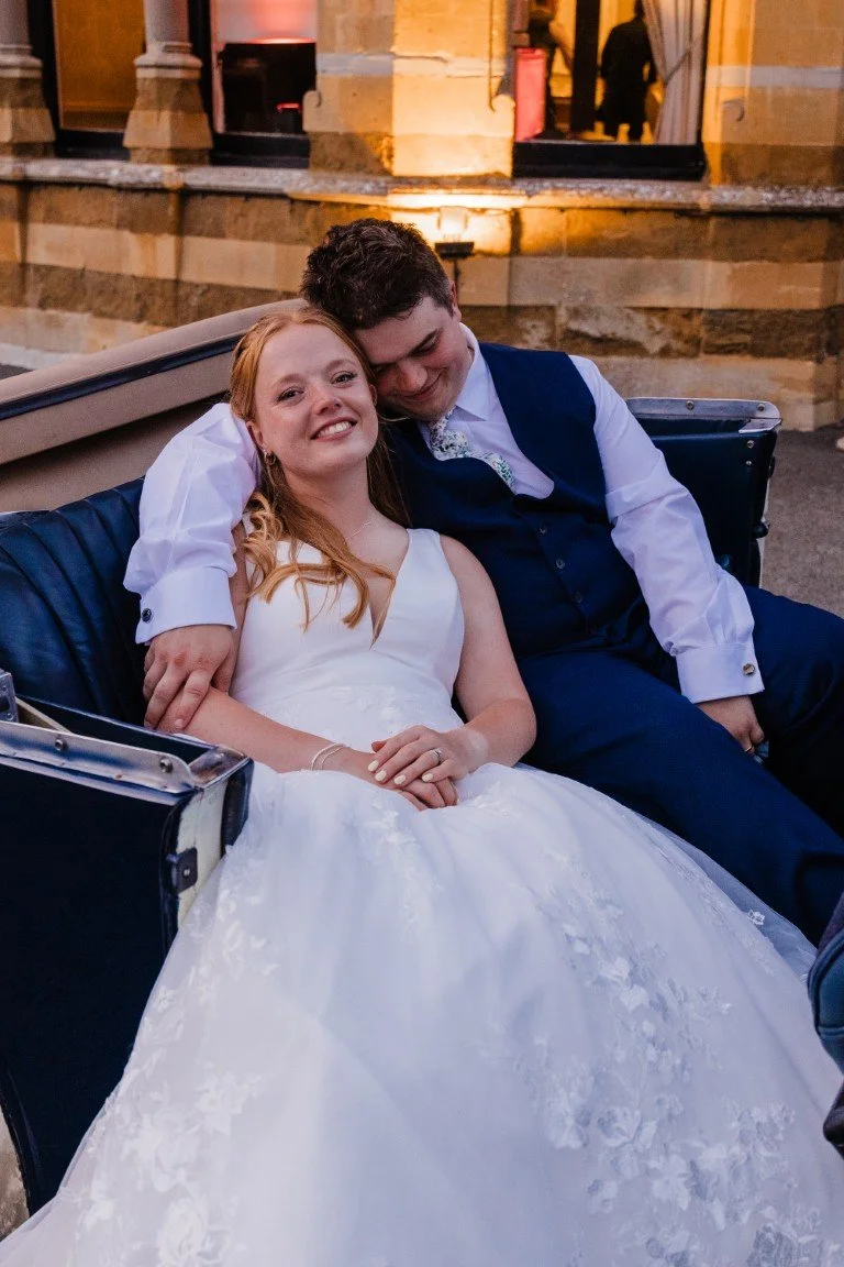 A newlywed couple in wedding attire sitting on a vintage car, smiling and relaxed, outside a building with warm lighting.