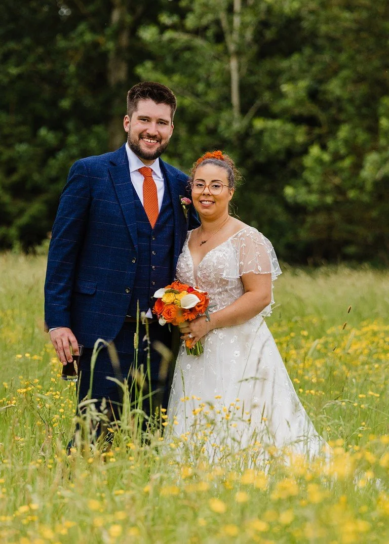 A newlywed couple standing in a field of yellow flowers with trees in the background, smiling at the camera. The groom is dressed in a blue plaid suit with an orange tie, and the bride is wearing a white lace wedding dress holding a bouquet of orange
