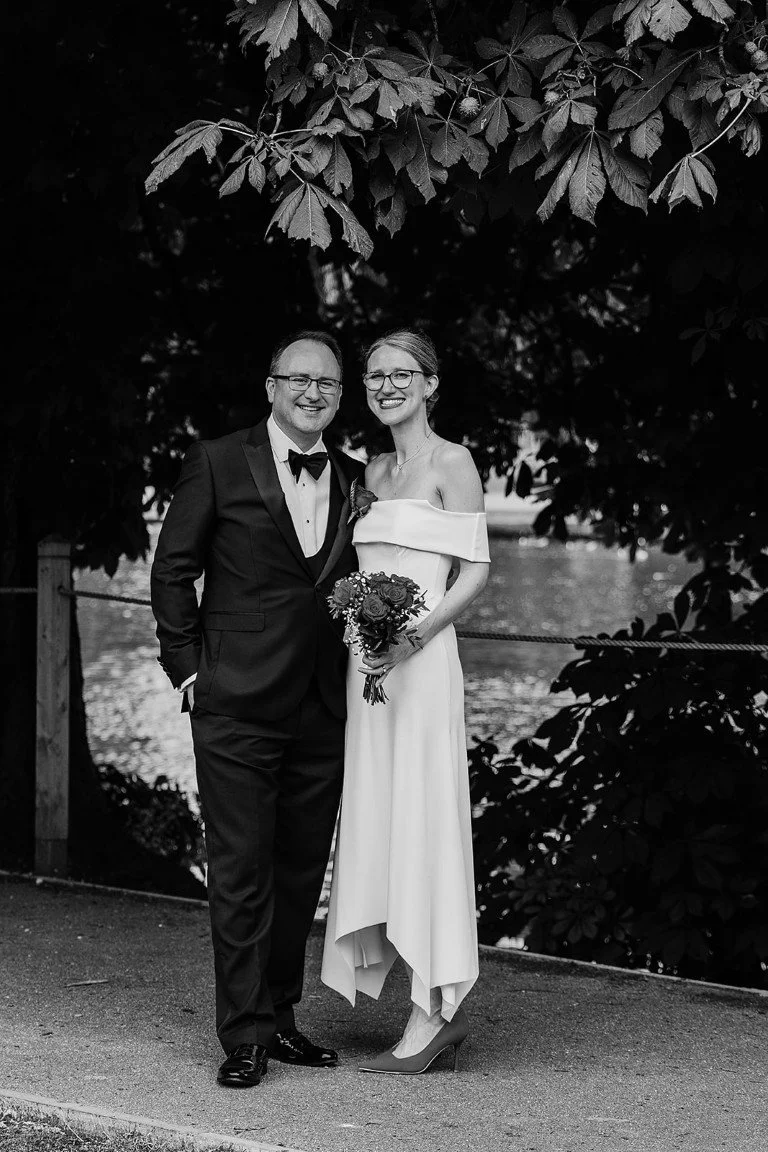 Black and white photo of a man and woman dressed in wedding attire, standing outdoors under leafy trees by a body of water, smiling at the camera. The man wears a tuxedo and the woman wears an off-shoulder dress and holds a bouquet.