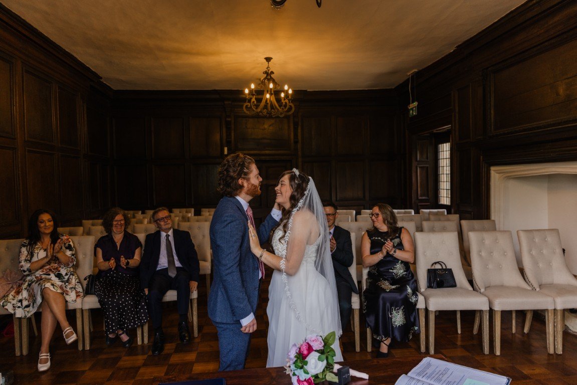 A bride and groom holding hands and smiling at each other during their wedding ceremony in a wood-paneled room, with six seated guests clapping and smiling in the background.
