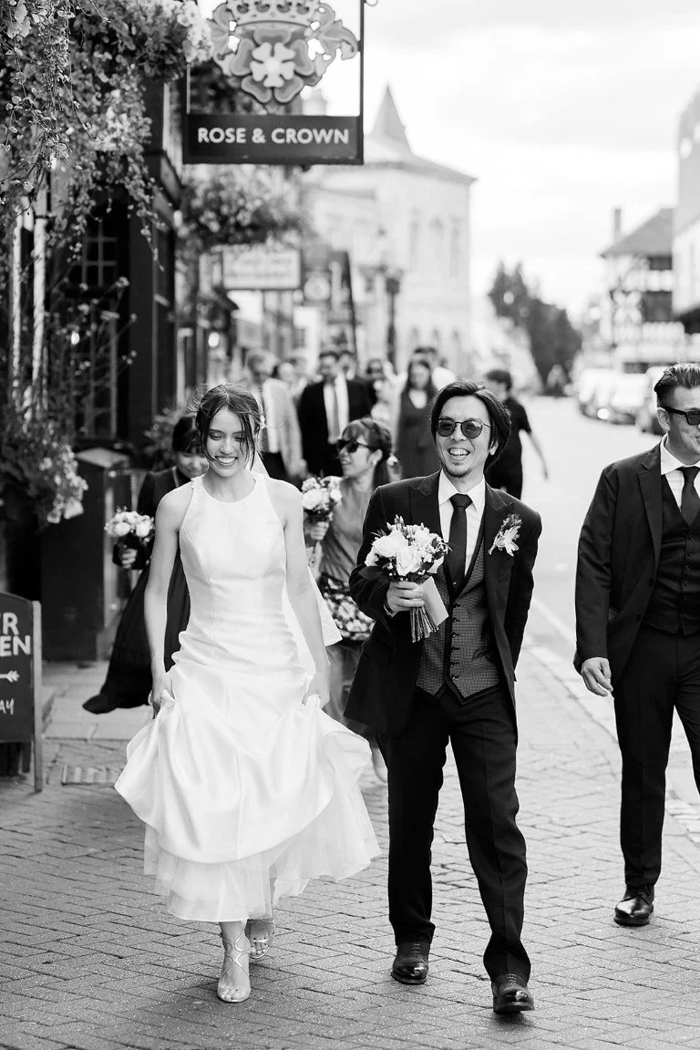 Black and white photo of a bride and groom walking on a city street, smiling, with friends around them, holding flowers, outside a venue named 'Rose & Crown'.
