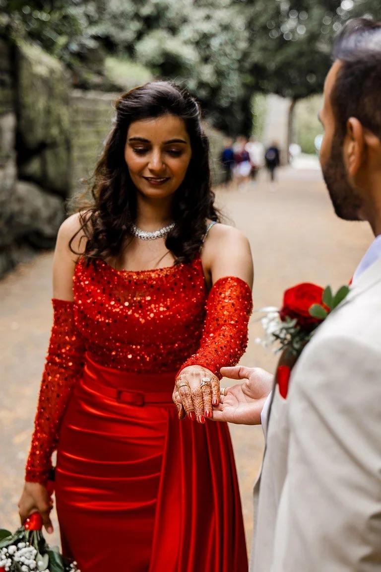 A woman in a red dress with sequined top and long sleeves showing henna on her hand, standing outdoors with a man in white suit holding her hand, holding a bouquet of flowers, with a blurred background of trees and people.