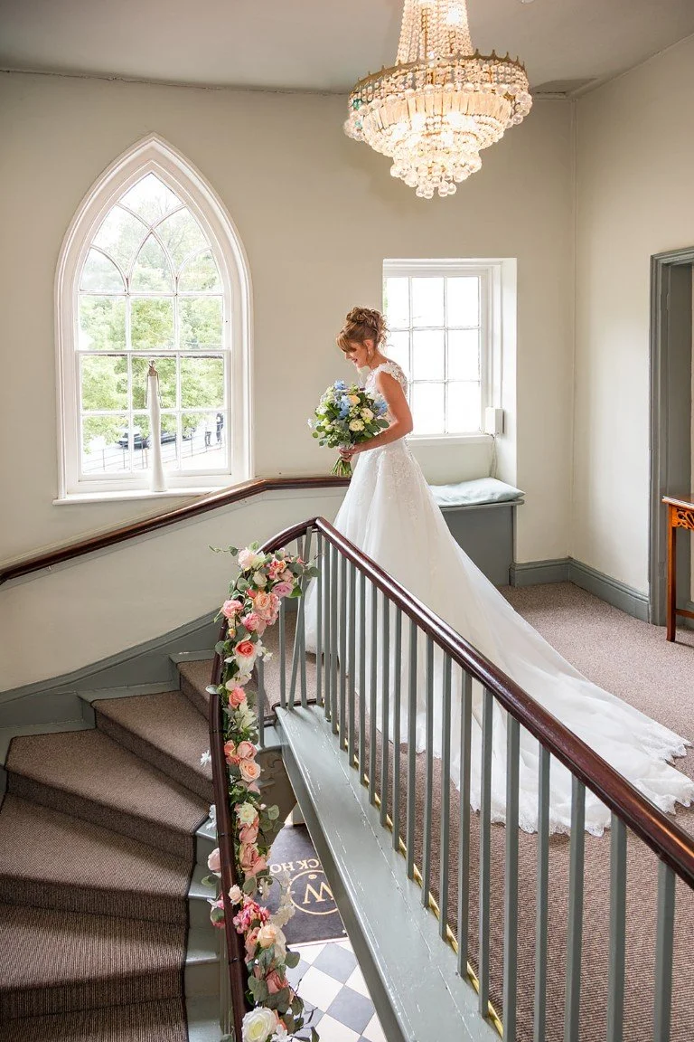 A bride in a white wedding gown with a long train standing on a staircase decorated with pink and white flowers. She is holding a bouquet of blue and white flowers, gazing down, in a room with a large arched window, a chandelier, and soft lighting.