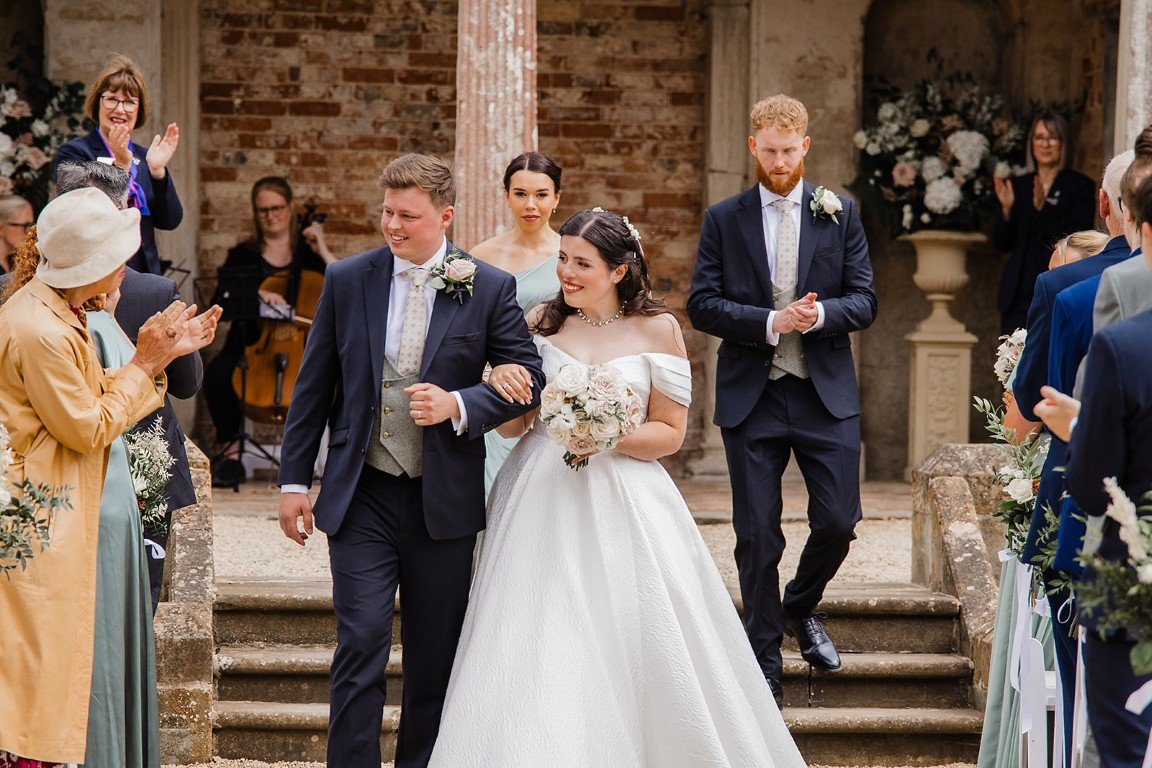 A bride walking down the aisle in a wedding ceremony, holding a bouquet, with groom and groomsman on either side, surrounded by seated guests and a small band in the background.