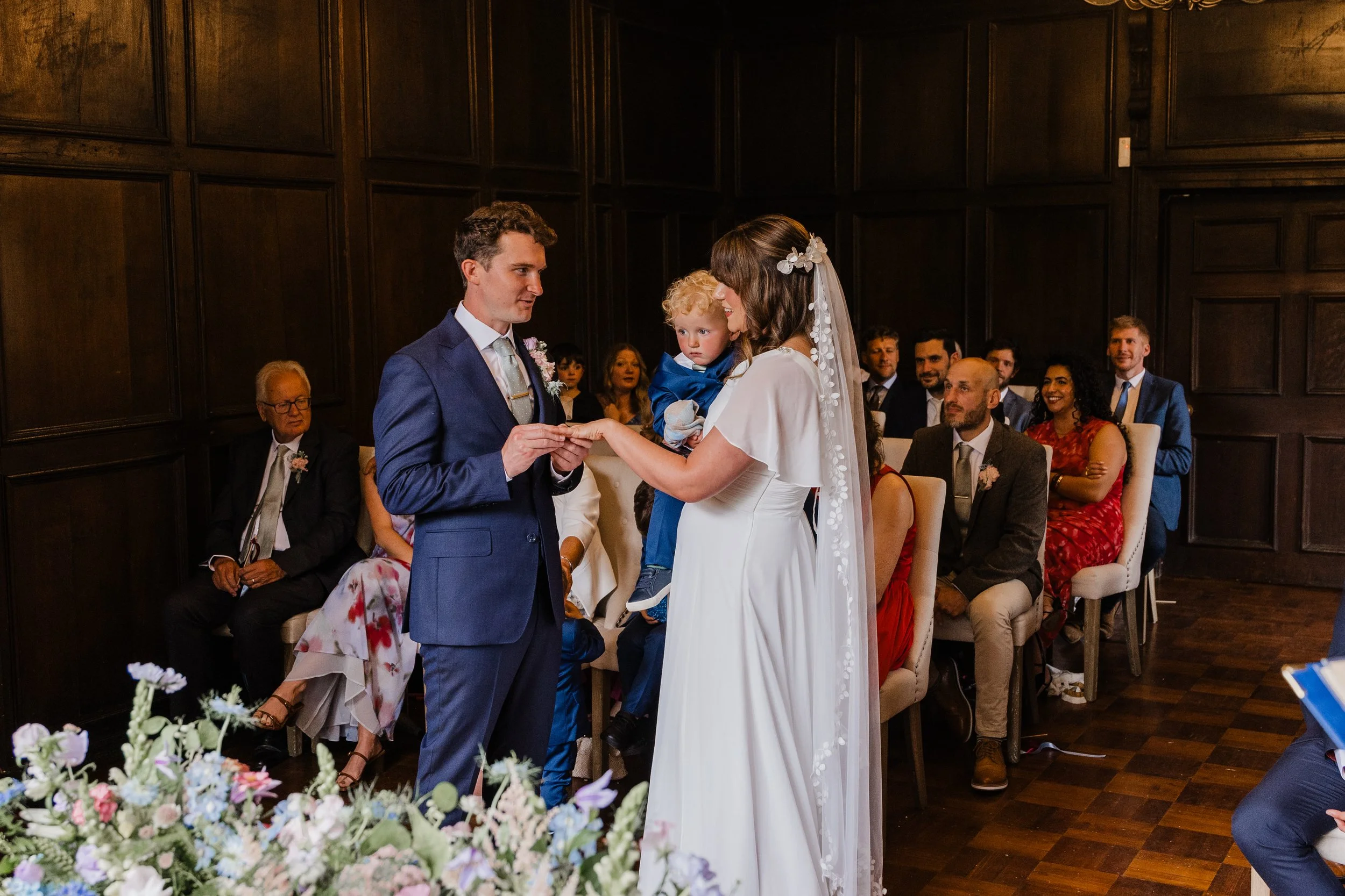 Bride and groom exchanging rings during a wedding ceremony, with guests seated in the background.