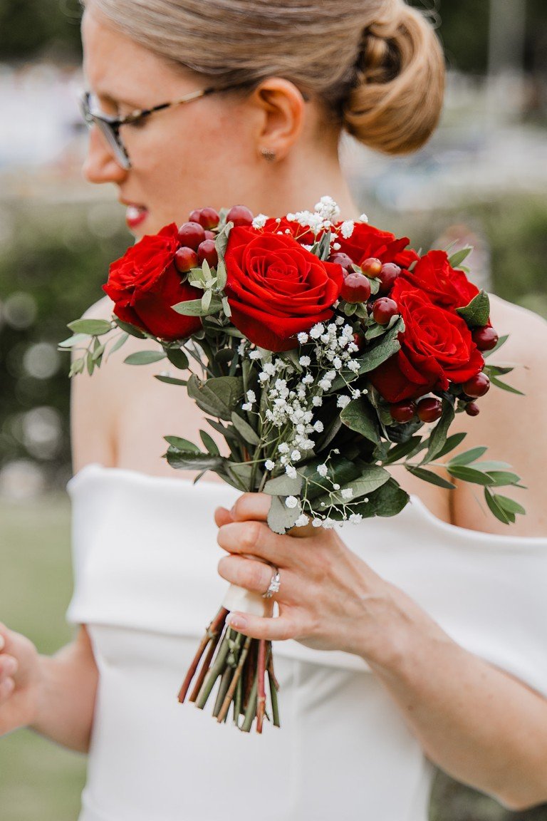 A woman in a white dress holding a bouquet of red roses, baby's breath, and greenery, with her face turned sideways and glasses on, outdoors.