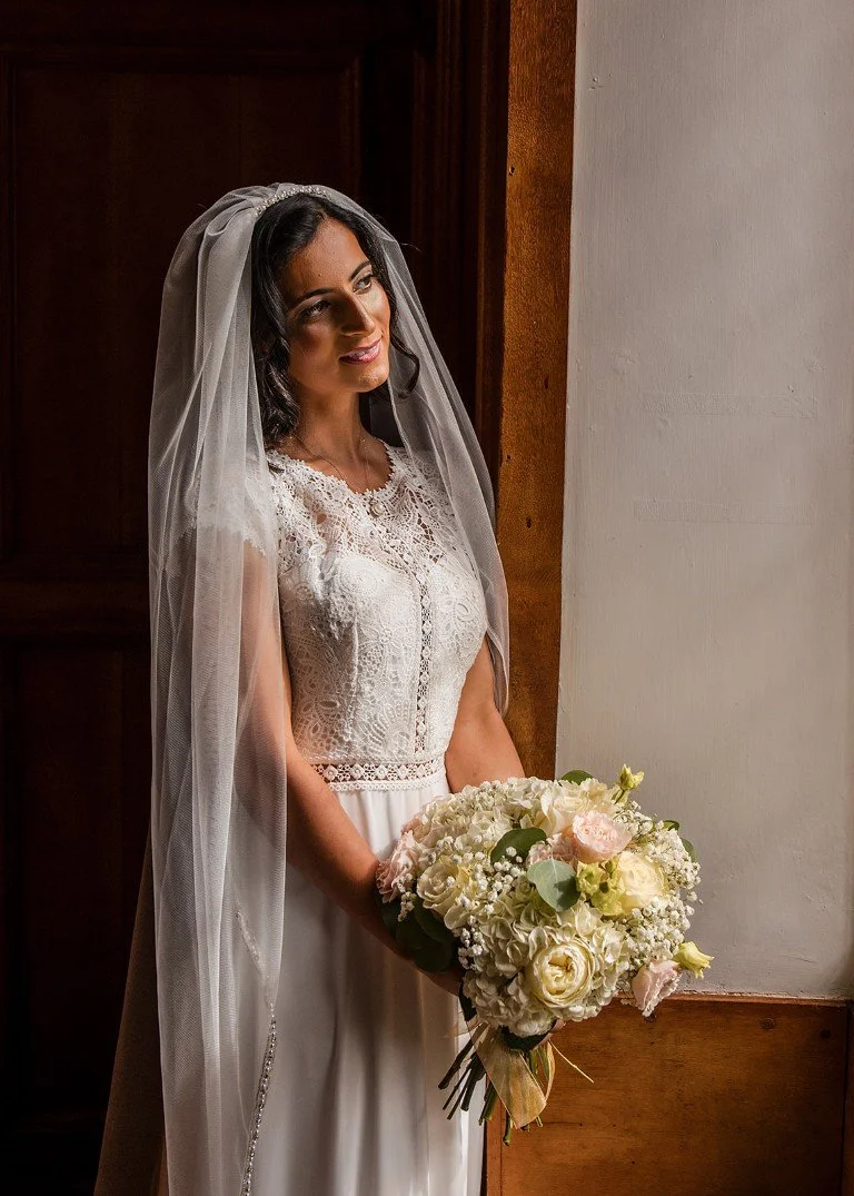 Bride in a lace wedding dress holding a bouquet of white and light pink flowers, with a veil on her dark hair, standing near a wooden door and white wall.