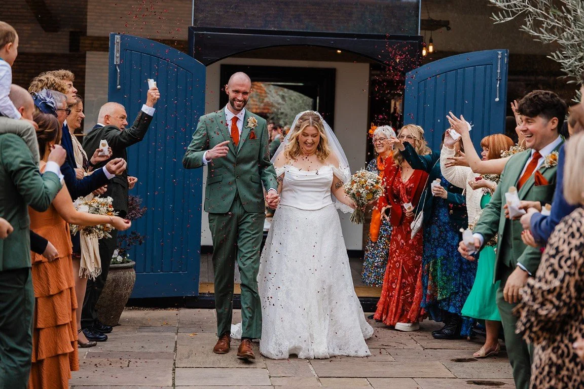 A bride and groom holding hands and smiling as they walk through a crowd of wedding guests who are cheering and throwing confetti outside a building with blue doors.
