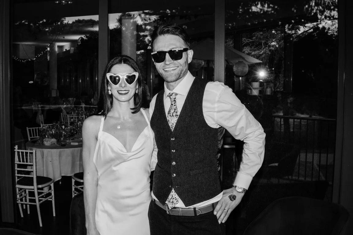 A man and woman dressed in formal attire at an indoor event. The woman is wearing a satin dress and heart-shaped sunglasses, while the man is dressed in a shirt, tie, and vest with sunglasses. They are smiling and standing close together.