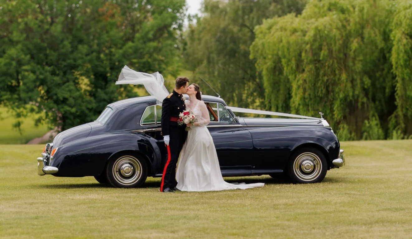 A bride and groom kissing in front of a black vintage car on a grassy field with trees in the background.