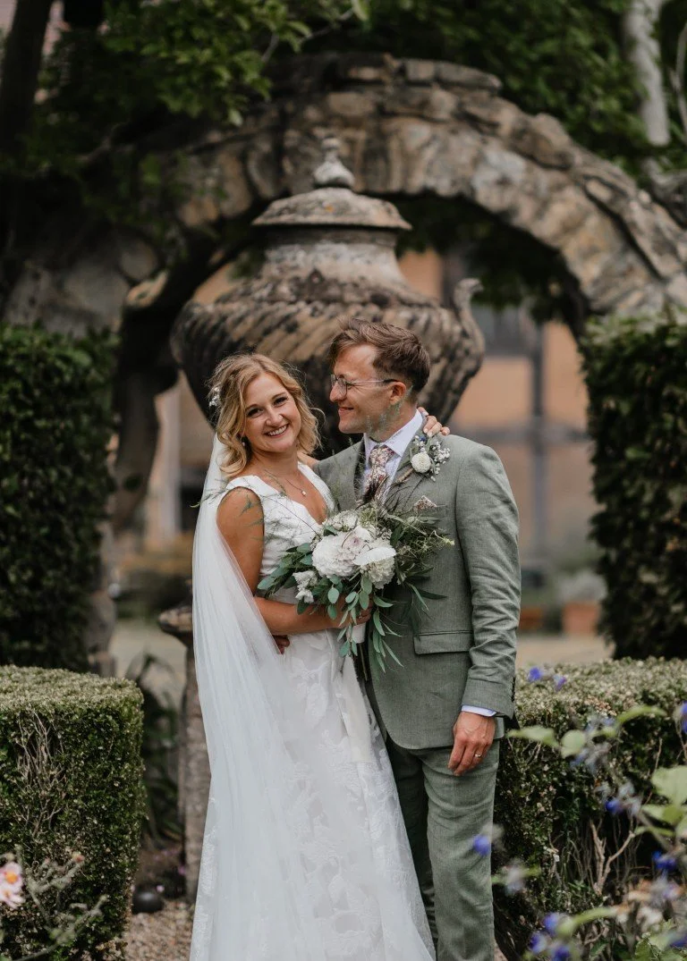 A bride and groom smiling and posing together outdoors in a garden setting for a wedding photo, with greenery and a decorative stone fountain in the background.