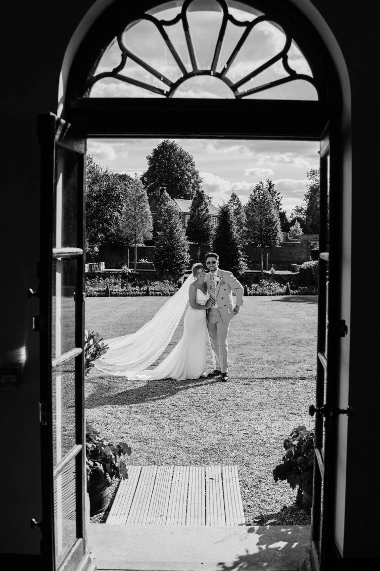 A bride and groom standing outdoors in a garden, viewed through an open window, with trees and a partly cloudy sky in the background.
