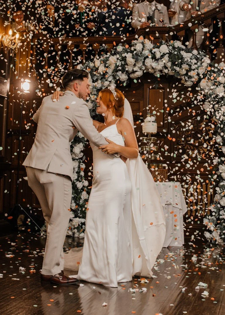 A couple is dancing and embracing at their wedding reception, surrounded by falling confetti and floral decorations, with guests watching from above.