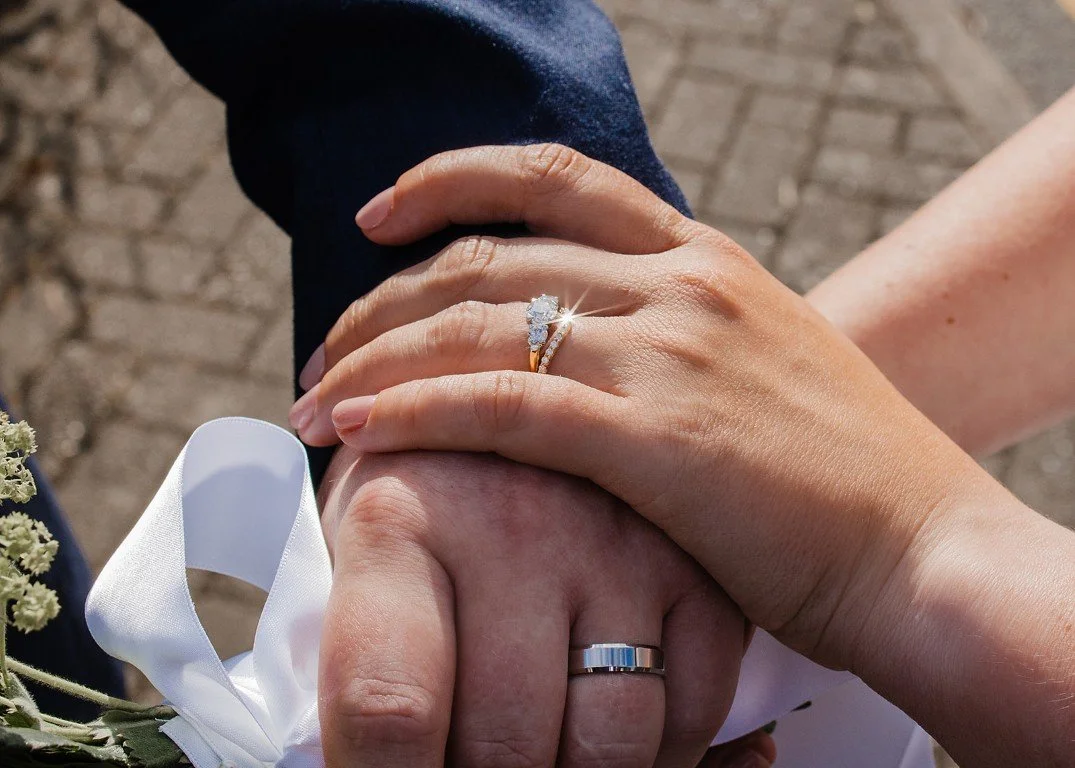 Close-up of a man and woman holding hands, with wedding rings on their fingers. The woman's hand has an engagement ring with a large diamond. The background shows a brick pavement.