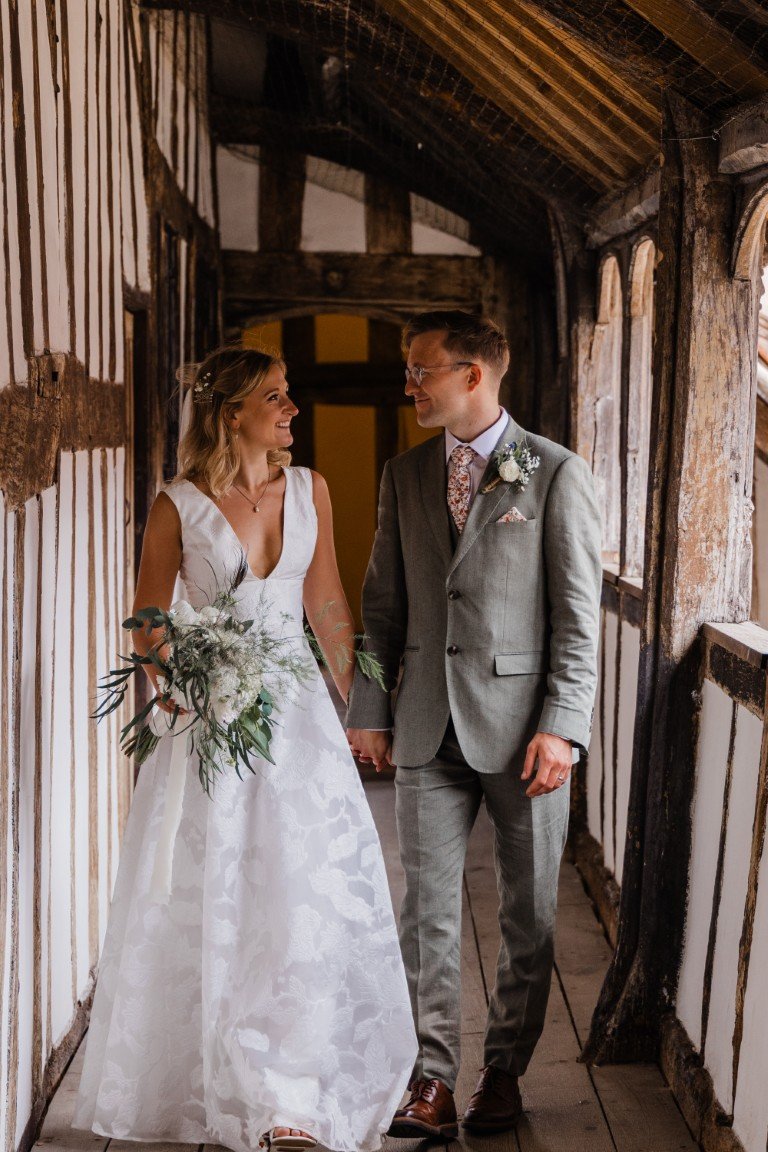 A bride and groom holding hands, walking through a rustic wooden hallway, smiling at each other on their wedding day.