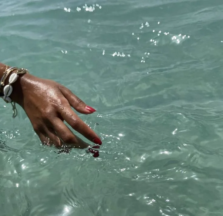A hand with red nail polish and a shell bracelet dipping fingertips into clear turquoise water.