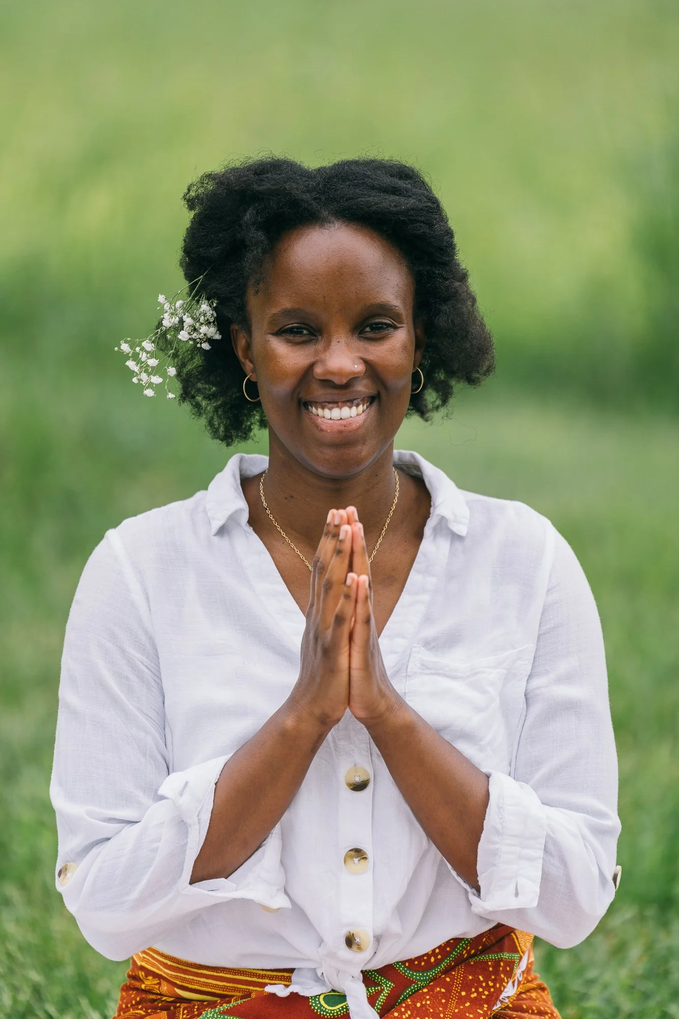 A woman standing outdoors with her hands pressed together in a prayer gesture, smiling at the camera. She has a flower tucked behind her ear, wearing hoop earrings, a white button-up shirt, and a colorful patterned skirt or sash.