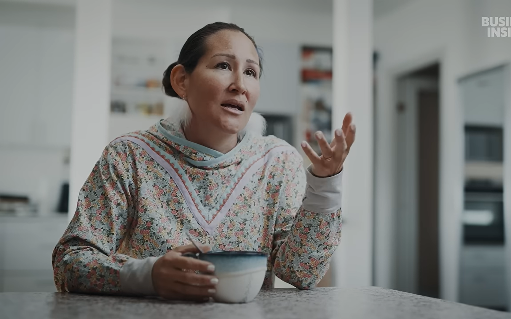 Qupanuk Olsen in her home speaking to the camera holding a cup of tea and wearing inuit-inspired earrings and clothing.