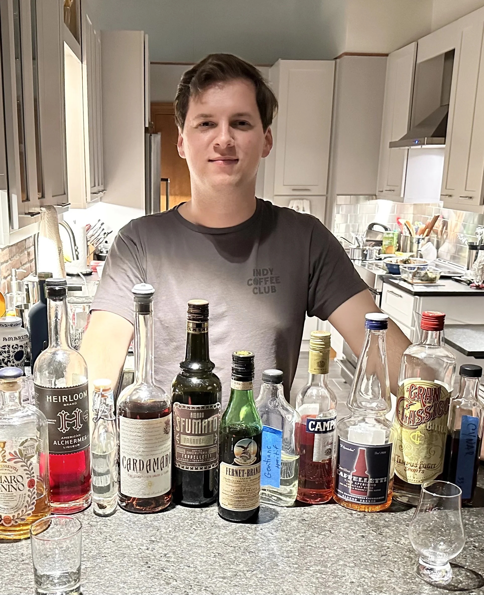 A man standing behind a kitchen counter with various bottles of alcohol in front of him.