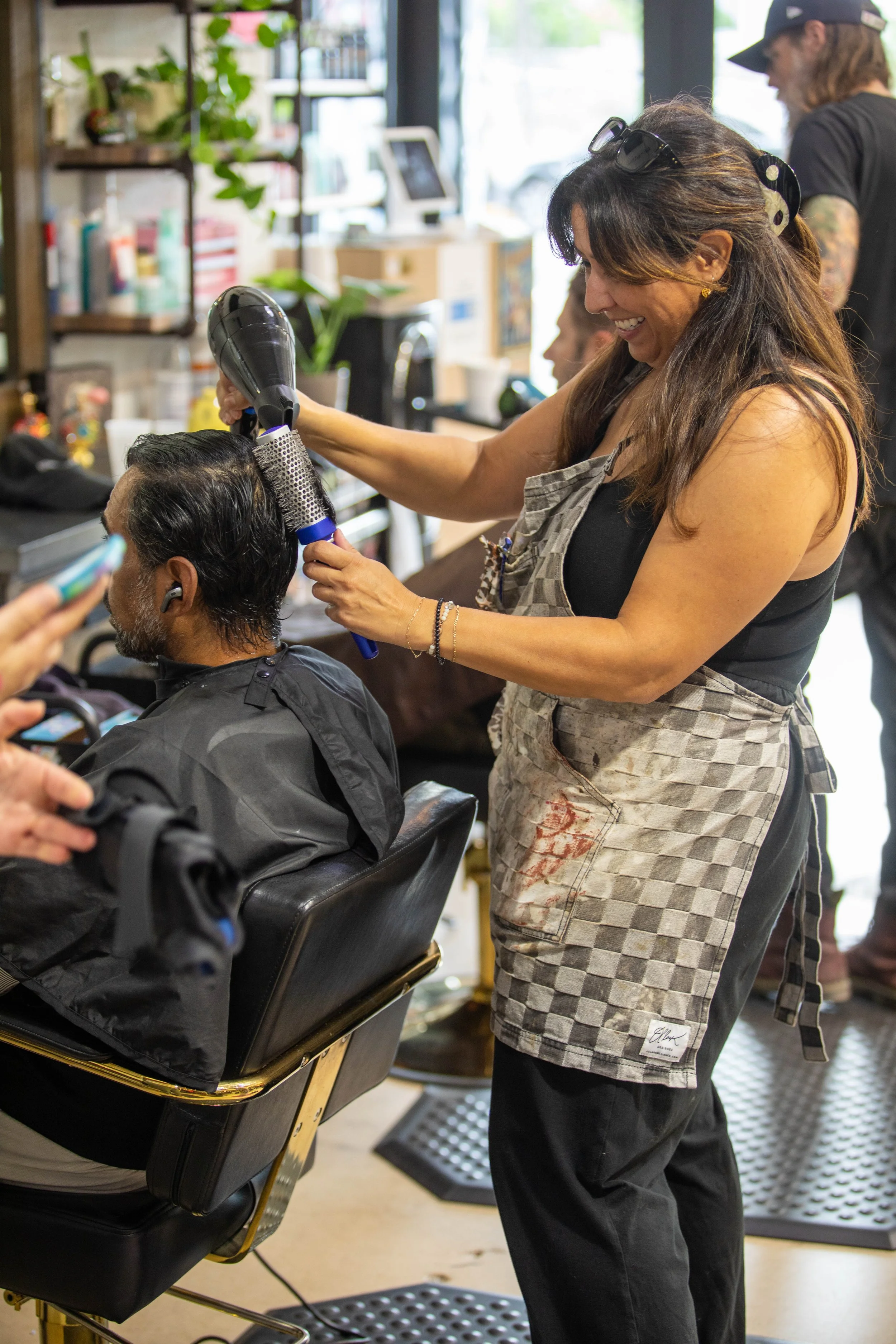 A woman with brown hair, wearing a checkered apron, is blow-drying a man's hair in a barber shop. The man is seated in a barber chair, and other people can be seen in the background.