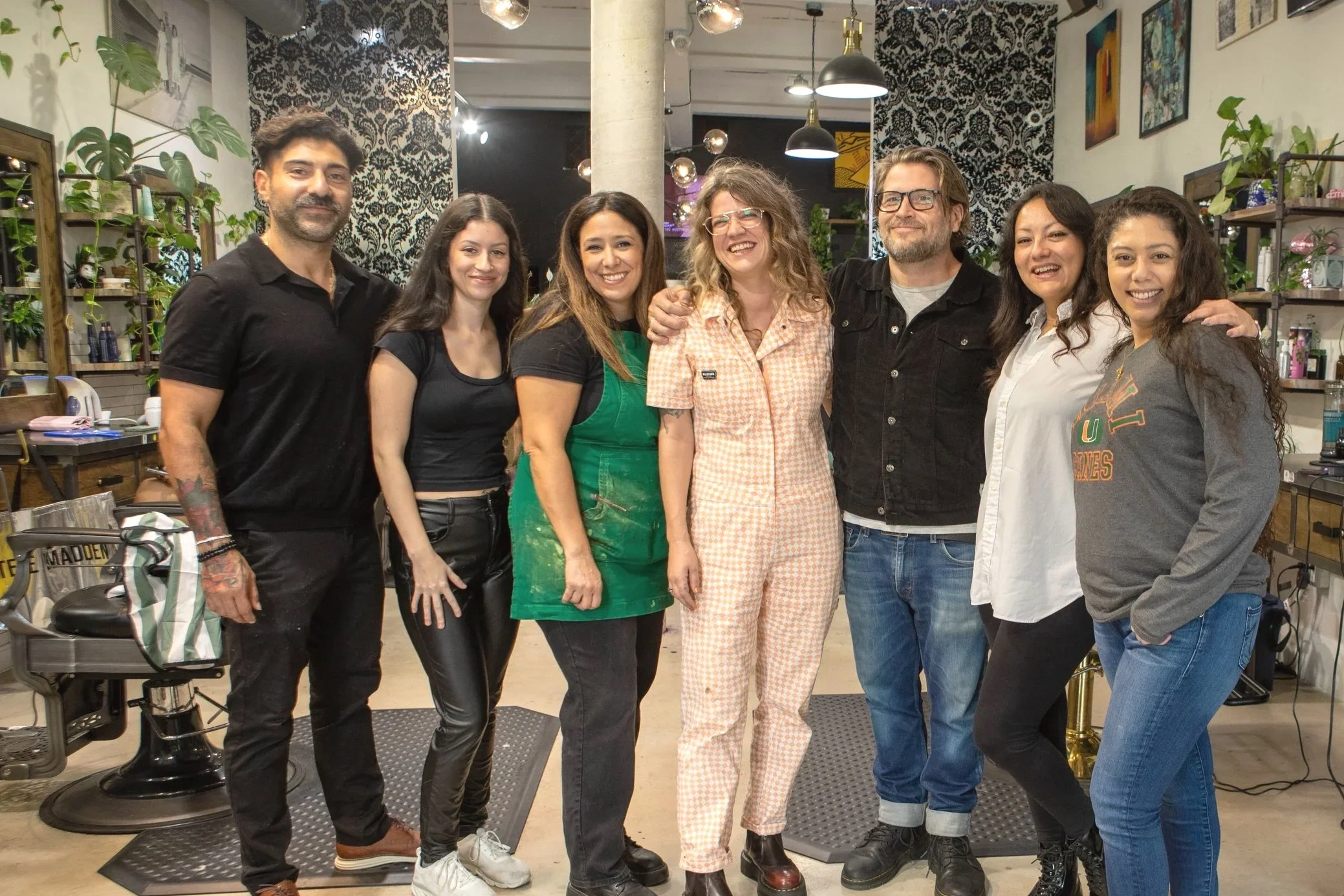Group of eight people smiling and standing together inside a stylish, plant-filled room with black and white patterned wallpaper.
