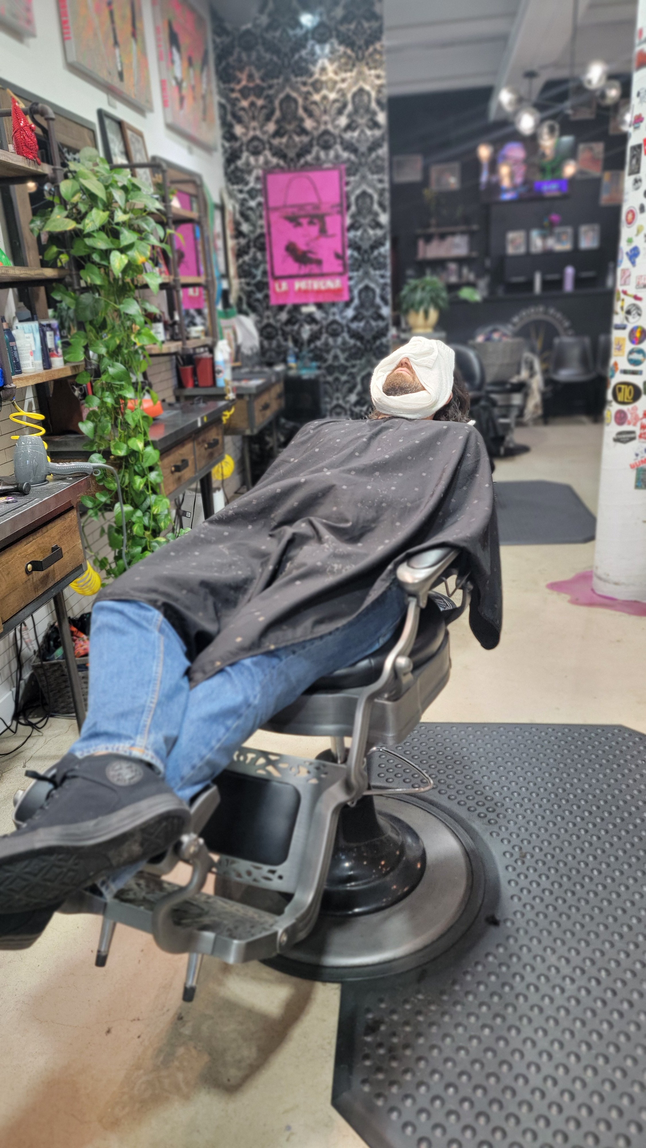 Man lying in a barber's chair with his eyes covered, wearing a fun black cape, jeans, and sneakers inside a trendy barbershop or salon.
